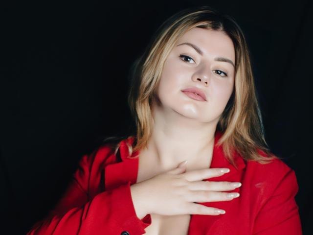 ZannaDreil with blonde hair, red shirt, and pale skin, has a hand on her chest, looking slightly up with a neutral expression, against a dark background.