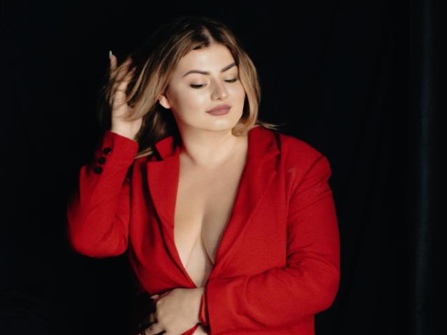ZannaDreil with blonde hair, wearing a red blazer, has her hand in her hair, exposing her tits, with a slight smile, against a dark background.
