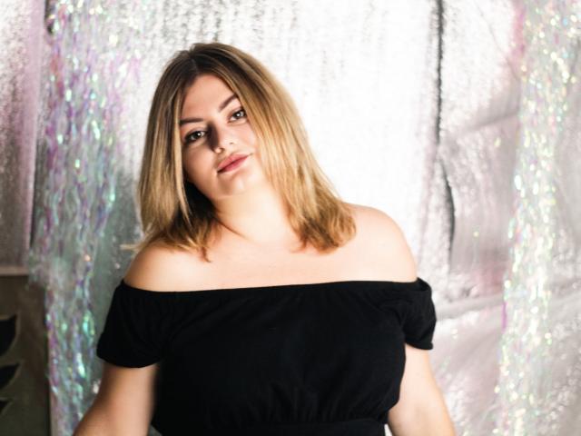 ZannaDreil with shoulderless black top, blonde hair, and slight smile, showing her tits and soft, curvy ass in the photo. She's looking slightly to the side, relaxed.