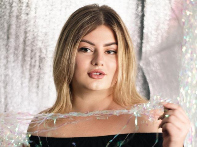 ZannaDreil with blonde hair, wearing a black off-shoulder top, holding iridescent silver strands in front of her chest, looking slightly to the side with a neutral expression.