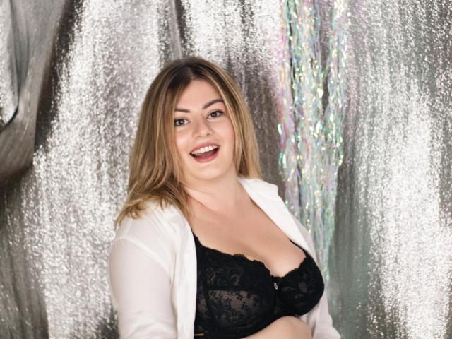 ZannaDreil with medium tits in a black lace bra, smiling, wearing an open white shirt, has blonde hair, and a cute face, in a silver textured background.