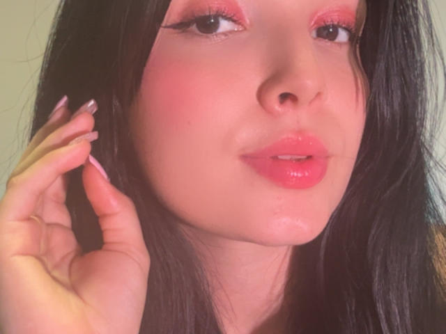 El rostro de Sarah Good, con sombra de ojos rosa, labios rosados brillantes y piel con un aspecto húmedo y brillante. Está tocándose el cabello negro con uñas pintadas de rosa, mirando ligeramente hacia un lado..