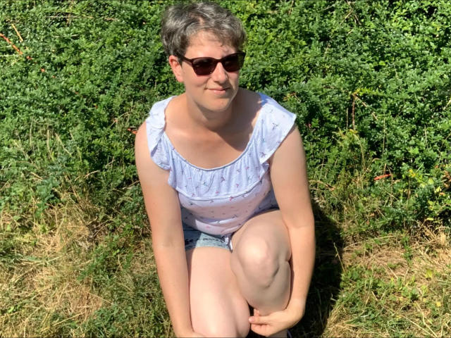 Milf01-hot with short gray hair, wearing sunglasses, a light blue ruffled top, and denim shorts, squatting on grassy ground, showing her soft ass and light skin.