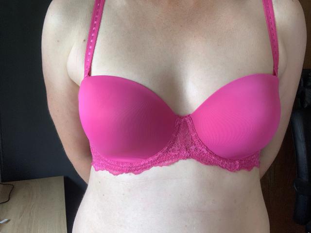 Milf01-hot is wearing a bright pink, lace-trimmed bra that highlights her full, round tits. Her fair skin contrasts with the vibrant bra. She's standing with arms behind.