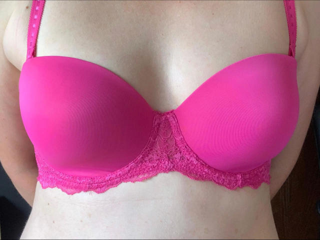 Milf01-hot's pink bra-covered tits, smooth and round, with lace underband, showing a slight tan line on her pale skin. The bra is bright pink and slightly padded.