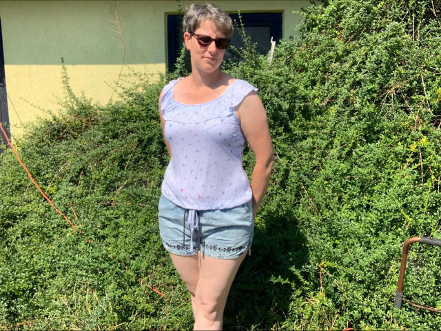 Milf01-hot with short gray hair, wearing sunglasses, a white patterned top, and denim shorts, standing in front of green shrubbery. She's got a small ass and a cute face.