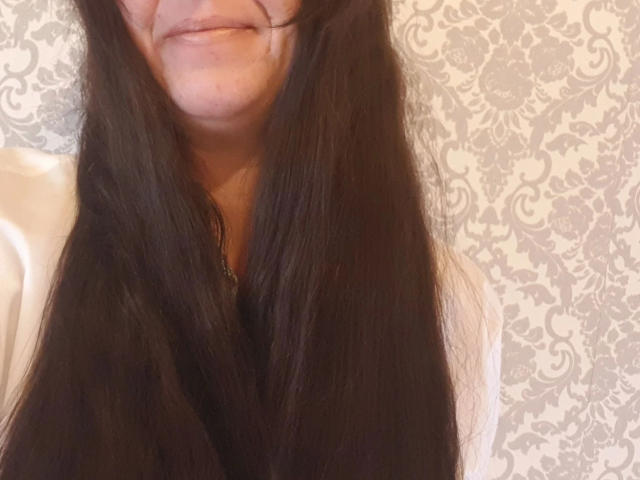Mistress-lola-hot with long black hair covering her tits, smiling slightly, in a photo showing just her upper body and face. Her ass is not visible.