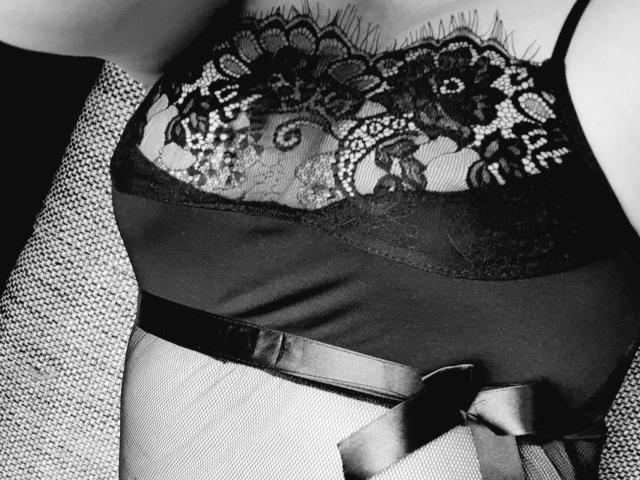 Mistress-lola-hot's black lace bralette with floral patterns, sheer fabric, and satin belt, showing her tits and cleavage, in a black-and-white photo.