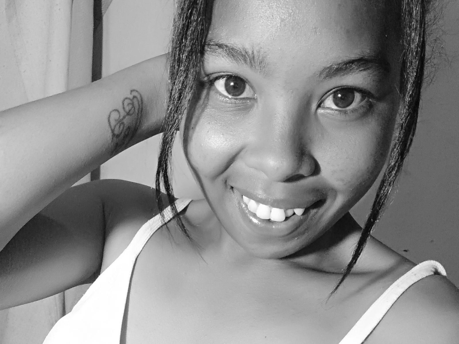RallyAnah69 has dark skin, a heart tattoo on her arm, and is smiling with her teeth showing. She's wearing a white tank top and has her hair in braids.