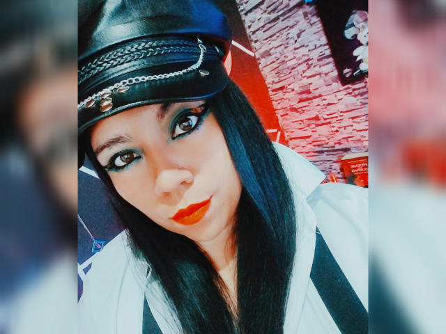 LexyDom with long black hair, wearing a black leather cap with chain, white shirt, and red lipstick, has bold eyeliner and red lipstick, looking directly at the camera.
