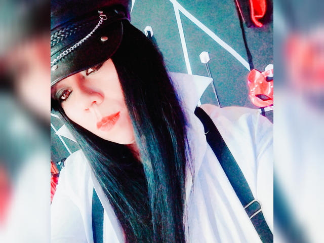 LexyDom with long black hair, wearing a black hat and white shirt with black suspenders, has pouty red lips and a slightly serious expression. Her tits are partially visible.
