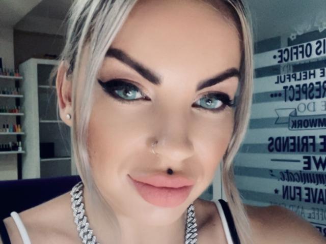 GoddessAlice has blonde hair, blue eyes, black eyeliner, full pink lips, a nose piercing, and a septum piercing, wearing a silver necklace, looking directly at the camera.