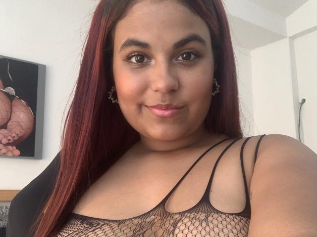 ValeenttiinaHot with red hair, brown eyes, and pink lips, wearing a black mesh top, showing her tits, smiling softly, and wearing small, gold, heart-shaped earrings.