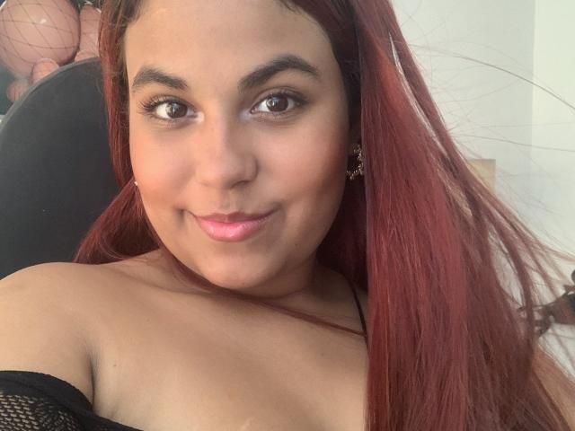 ValeenttiinaHot with red hair, brown eyes, and pink lipstick, smiling at the camera, showing off her bare shoulders and black mesh off-shoulder top. She's got cute earrings too.