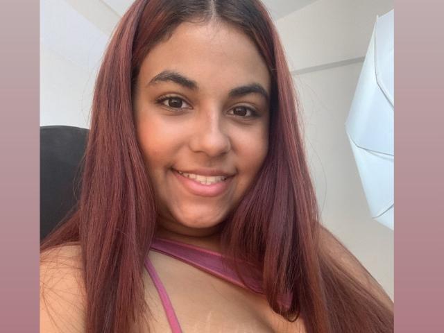 ValeenttiinaHot has long red hair, brown eyes, and a cute smile showing her pink lipstick. She's wearing a pink top, and her small tits are visible.