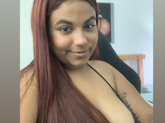 ValeenttiinaHot with red hair, brown skin, and small tits, smiling slightly, wearing a black spaghetti strap, showing a tattoo on her right arm.