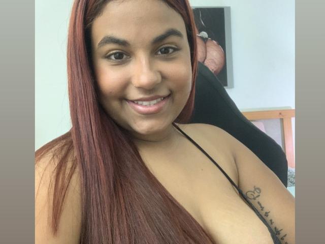 ValeenttiinaHot with long red hair, smiling, tan skin, black strap top, showing a tattoo on her right arm, and medium-sized tits. Background includes a blurred, seated man.