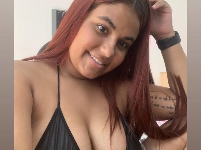 ValeenttiinaHot with red hair, smiling, wearing a black halter top, showing her large tits, tattoo on right arm, black wristband, looking at camera.