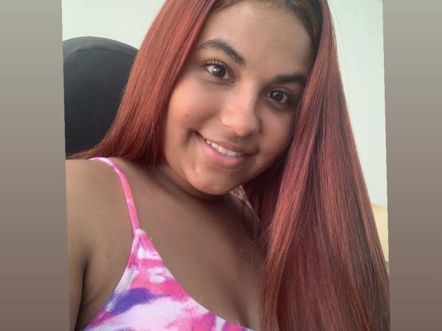 ValeenttiinaHot with long red hair, brown eyes, and a smile, wearing a pink and purple tie-dye top, showing small tits and a cute face.