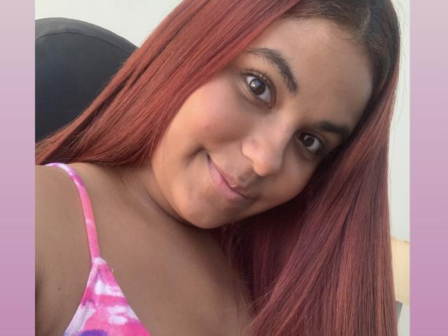 ValeenttiinaHot with red hair, brown eyes, and a slight smile, wearing a pink tie-dye top, showing her cute face and small nose. Her tits are not visible.