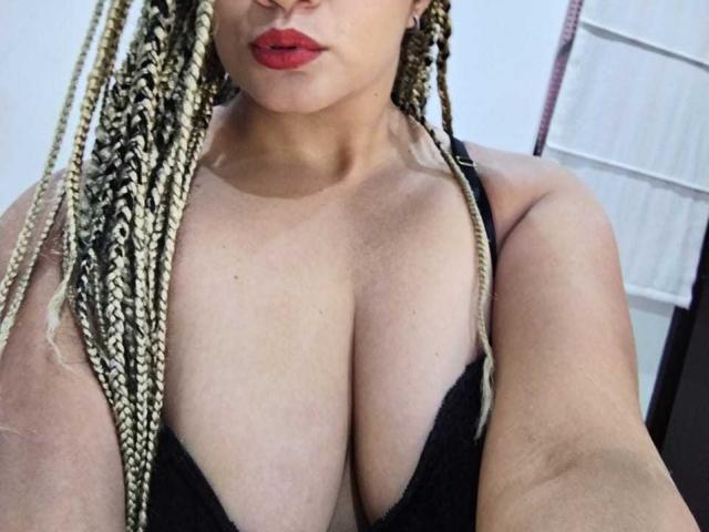 ViolettaChaudeX mit blonden Zöpfen, roten Lippen und großen Brüsten in einem schwarzen Top, wobei ihre Brust und ihr Dekolleté auf dem Selfie deutlich sichtbar sind..