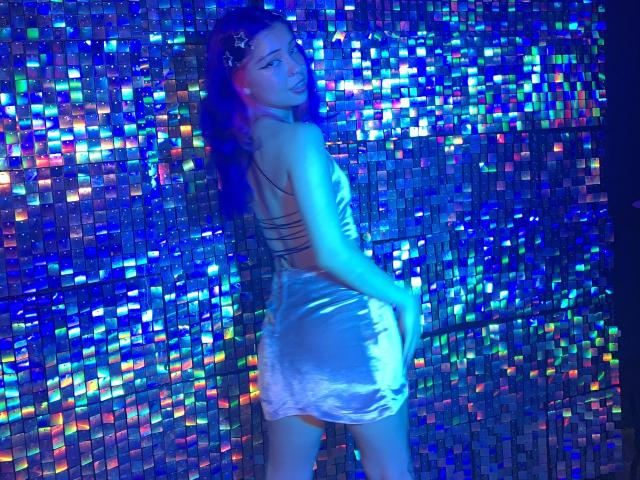 LiaRose69 in a shiny silver dress, back turned, hand on her ass, looking over her shoulder, blue neon lights reflecting on her skin and dress.