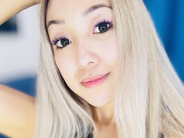 RenyLime has blonde hair, blue eyes with purple eyeliner, and pink lipstick, smiling with a slight blush, arms raised behind her head, looking cute and attractive.