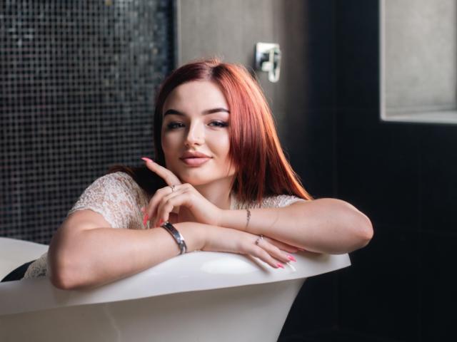 NicolPots with red hair, white lace top, pink nails, finger pointing up, smiling, leaning on white bathtub, black tiled wall, silver bracelet, pink lipstick, light skin, sexy.