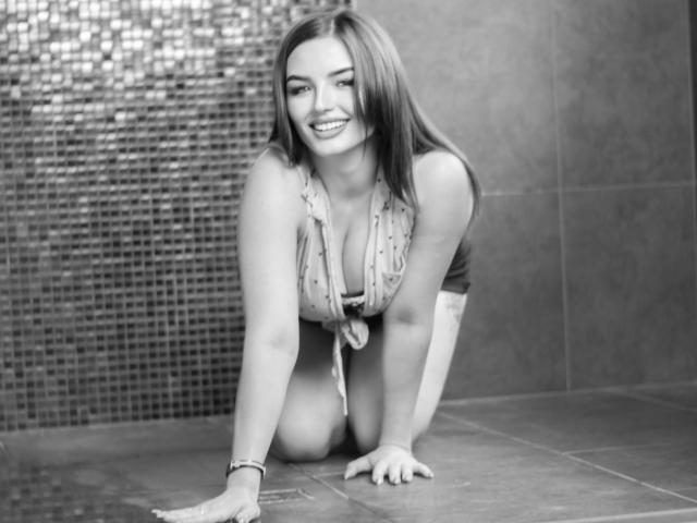 NicolPots on all fours, smiling, with medium tits, wearing a tied-up top, in a tiled room, looking happy, right hand on the floor, left hand in front.