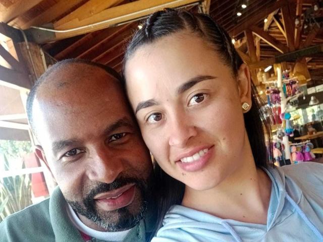 IdalyAndCarlos, a cute brown-skinned guy with a beard, and a white-skinned woman with braids, smiling and leaning in for a selfie, both showing happy, relaxed faces.