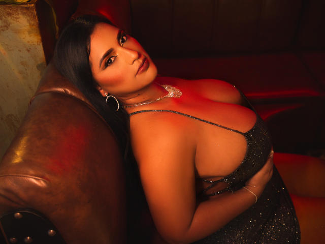 KorinaKova with big boobs, wearing a sparkly black dress, sitting on a brown leather couch, red light on her, looking at the camera with a sultry expression.