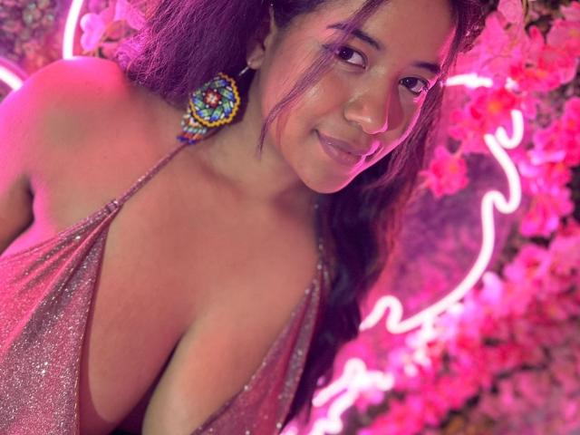 AllisonLovS with medium brown skin, long purple hair, wearing a sparkly pink halter top that reveals her big, shiny tits. She's smiling, wearing colorful earrings, neon lights behind.