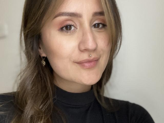 NinaBryons has brown eyes, a small nose piercing, and a gentle smile. She's wearing a black turtleneck, with wavy brown hair and small gold earrings.
