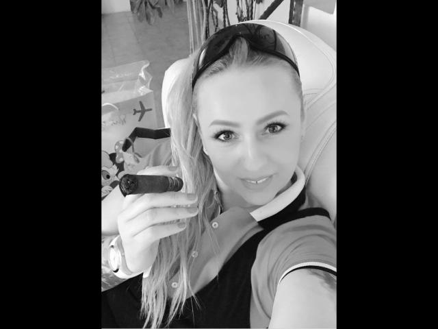 Black and white selfie of krystal75-hot with long blonde hair, wearing a polo shirt, holding a cigar in her right hand, smiling, and wearing sunglasses on her head.