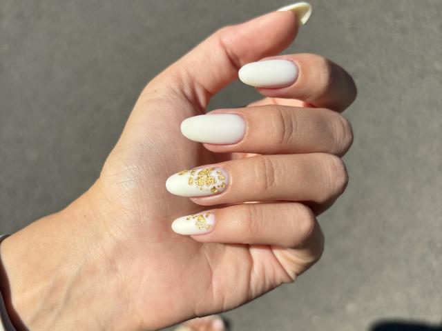 Pingli's hand with white, almond-shaped nails, gold glitter on two nails, sunlight, and a grey background. Boobs are not visible.