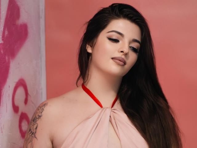 PamDares, com cabelos castanhos longos, vestido vermelho de alças, tatuagem no ombro esquerdo e maquiagem sedutora, olhando para baixo com os lábios levemente entreabertos, pele clara e maquiagem discreta..