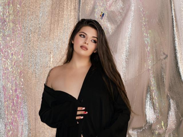 PamDares with long brown hair, dark makeup, and red nails, wearing a black off-shoulder shirt, exposing her tits, standing against a glittery, shimmering background.