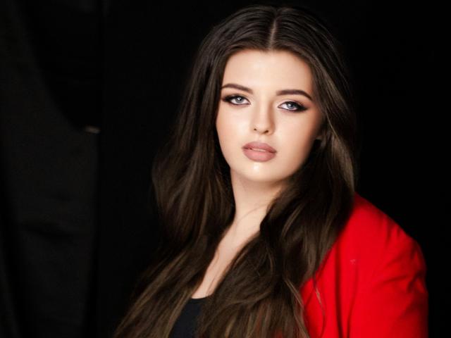 PamDares with long brown hair, blue eyes, and red jacket, looking at camera with slightly parted lips, wearing black top, against black background.