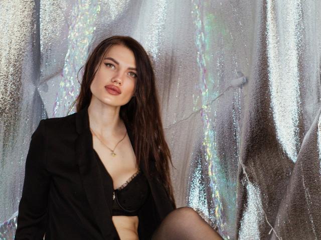 MilaLoona with long brown hair, black blazer, black lace bra, and black lace garter, showing her tits and leg with a tattoo, sitting against shiny silver fabric.
