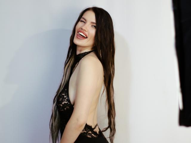MilaLoona with long dark hair, smiling, wearing black lace lingerie, showing her back and sideboob, standing against a white wall. Her ass is slightly visible from the side.