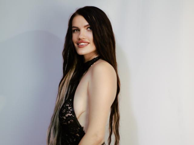 MilaLoona with long dark brown hair, smiling, wearing a black lace halter top, exposing her ass, standing sideways, showing her tits. She's fair-skinned with a cute face.