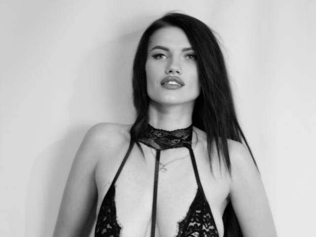 Black-and-white photo of MilaLoona with long dark hair, wearing a lace halter top that reveals her tits and a small tattoo on her chest. She's looking slightly to the side.