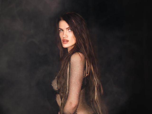 MilaLoona with long brown hair, wearing a sheer, gold mesh top, exposing her small tits, and looking over her shoulder with a serious expression in a dark, smoky photo.