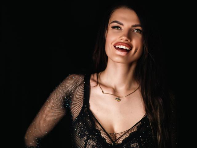 MilaLoona met donker, golvend haar, lachend, draagt een zwarte, doorschijnende, glinsterende top en een delicate ketting, waarbij haar borsten zichtbaar zijn, op een zwarte achtergrond..