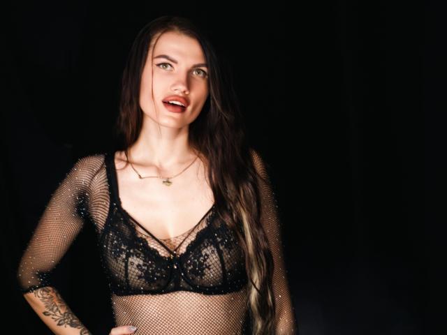 MilaLoona with long brown hair, wearing a black mesh top and lace bra, showing her tits, tattoo on left arm, necklace, dark background, smiling with red lipstick.