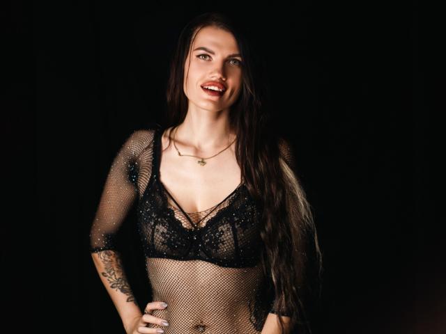 MilaLoona with long dark hair, smiling, wearing a black lace bra and sheer fishnet top, showing tattoos on her right arm and hand on dark background.