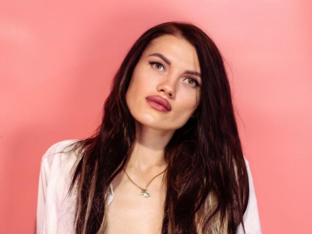 MilaLoona has long, dark brown hair, light skin, brown eyes, and red lipstick. She's wearing a white shirt and a gold necklace with a small, colorful pendant.