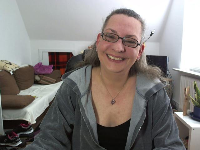 CashLadyVivian - attraktiv, lächelnd, mit Brille, grauer Kapuzenjacke, schwarzem Oberteil, Halskette, braunes Haar im Pferdeschwanz, sichtiger Hintern unter der Kapuzenjacke, weißer Raum, braunes Sofa, pinke Tasche im Hintergrund..