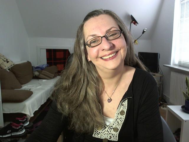 CashLadyVivian-hot with glasses, smiling, long brown hair, black cardigan, white lace top, necklace, medium tits, cute, attractive, sitting in a bright room with a window.
