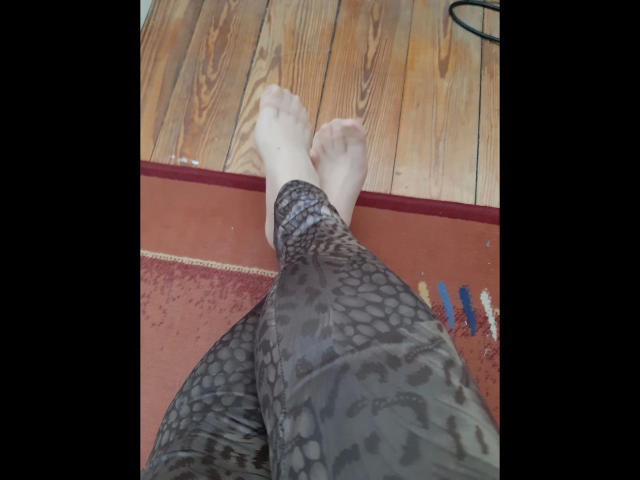 Las piernas de CashLadyVivian-hot, vestidas con pantalones de estampado de leopardo, con los pies descalzos cruzados sobre el suelo de madera. Sus pies apuntan hacia arriba, mostrando las plantas, mientras que sus piernas están ligeramente separadas..